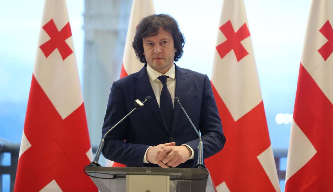 a man in a suit and tie stands at a podium with flags behind him
